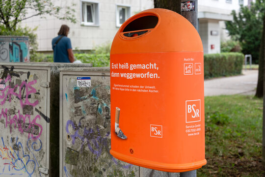 Berlin, Germany - 16 August 2026. Orange street cigarette bin mounted on a pole beside graffiti-covered utility boxes in an urban area. Urban waste management, smoking, environment, civic cleanliness.