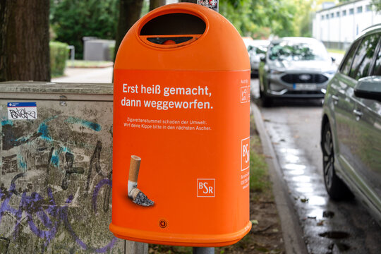 Berlin, Germany - 16 August 2026. Orange street cigarette bin mounted on a pole beside graffiti-covered utility boxes in an urban area. Urban waste management, smoking, environment, civic cleanliness.