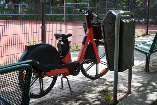 Berlin, Germany - 16 August 2026. Parked rental electric bike stands beside a trash bin at a fenced outdoor sports court. Urban mobility, sharing economy, transport, sustainability, city life.