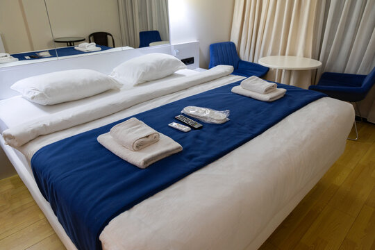 Interior view of a modern hotel room featuring a large double bed with fresh white linens, blue runner, neatly folded white bath towels, TV remote controls, and packaged disposable slippers placed on 