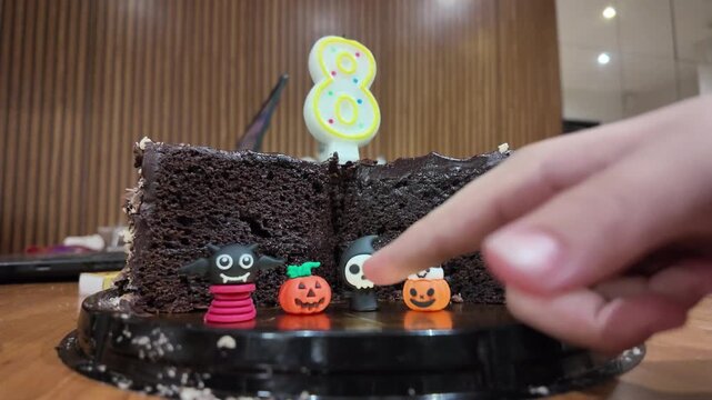 Hand points at chocolate cake with Halloween decorations. Yellow number eight candle stands tall. Small pumpkins and ghosts adorn the cake. Cake rests on a dark base with crumbs scattered