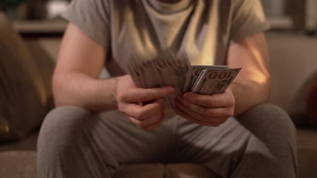 Man counts a thick stack of 100 US dollar bills while sitting at home. Hands flip through cash and banknotes. Money, income, savings, salary, wealth and personal finance concept.