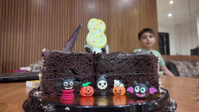 Chocolate cake with Halloween decorations. Child waves joyfully in background. Candle shaped like number eight. Small figures adorn cake surface. Warm indoor lighting enhances scene