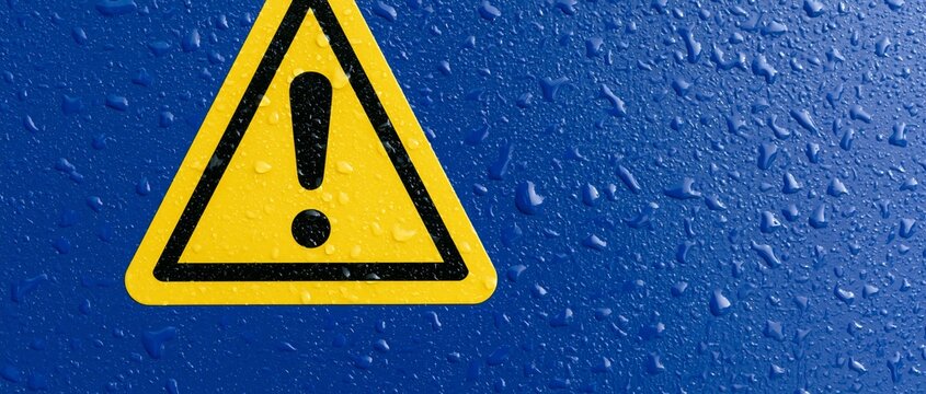 Yellow triangular warning sign with black exclamation mark mounted on wet vibrant blue painted wall covered in water droplets under bright overhead lighting