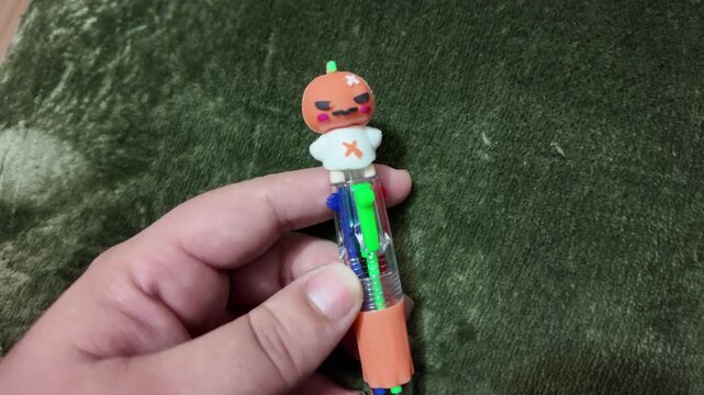 Hand holds colorful pen with pumpkin cap. Pen has bright colors and playful face. Green background adds cozy contrast. Person shows item with gentle touch. Perfect for festive or creative scenes