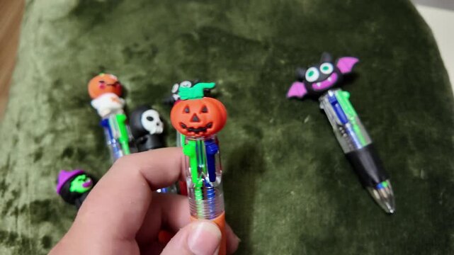 Hand holds colorful Halloween-themed pens. Pens feature pumpkins, bats, and ghosts. Green fabric provides soft, cozy backdrop. Childlike joy shown in playful selection