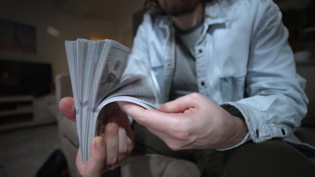 Man flips through a thick stack of cash while sitting at home. Hands hold many banknotes. Money, savings, income, salary, wealth, budget, payment and personal finance concept. Authentic lifestyle.