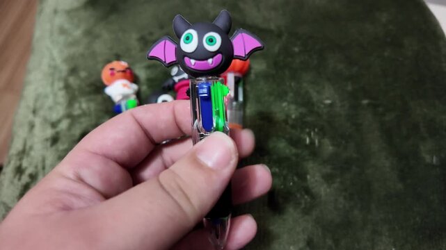 Hand holds colorful Halloween-themed pen with cartoon bat head. Soft green fabric background shows playful characters. Bright colors suggest fun, festive, or child-friendly use