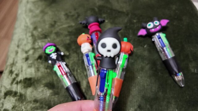 Hand holds colorful Halloween themed pens against green fabric. Pens feature pumpkins, bats, and skulls in bright, playful designs. Each pen is filled with multiple colored ink cartridges