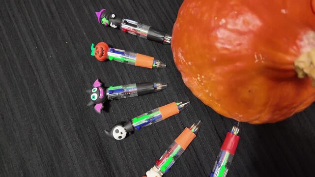 Halloween-themed pens scattered near a pumpkin. Black wood surface with orange pumpkin. Pens feature spooky faces and colors. Ideal for Halloween, seasonal marketing, or creative projects