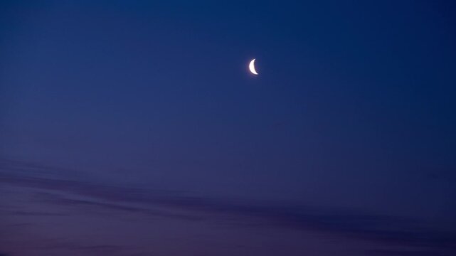 Time lapse of crescent moon rising against cloudless night sky above horizon.