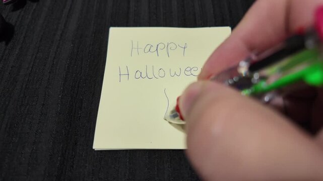 Hand writes "Happy Halloween" with a drawn ghost on sticky note. Person's hand guides pen to create a smiling, simple ghost. Black surface frames the note's festive, playful message