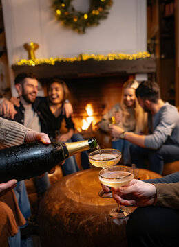 Group of close friends gathered in a cabin near the fireplace, having fun together celebrating Christmas or New Year 2026. Person pouring sparkling wine into glasses while laughing and embracing