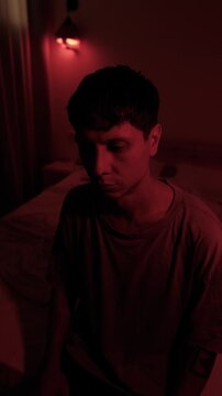 Anxious millennial man staring into the distance, lost in thoughts about life and uncertainty, portraying existential anxiety and emotional isolation in a dark red low key bedroom, vertical
