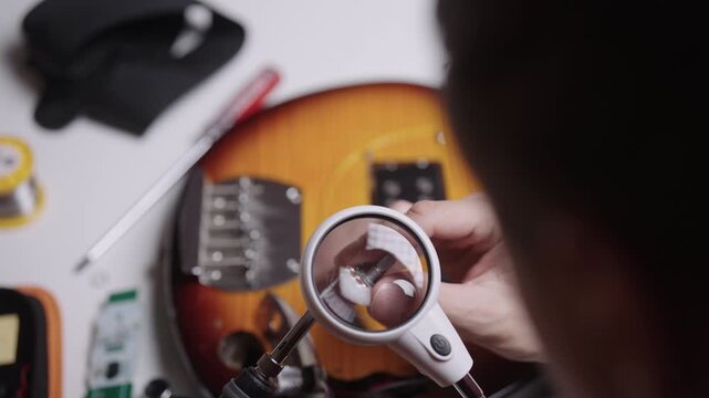 Over shoulder closeup of technician using magnifying glass to inspect guitar potentiometer. Electric guitar body and tools like solder and screwdriver on workbench, close-up, top view, slow motion.