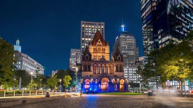 A wide angle timelapse of Trinity Church in Boston, Massachusetts, illuminated at night surrounded by tall modern skyscrapers under a deep blue twilight sky.