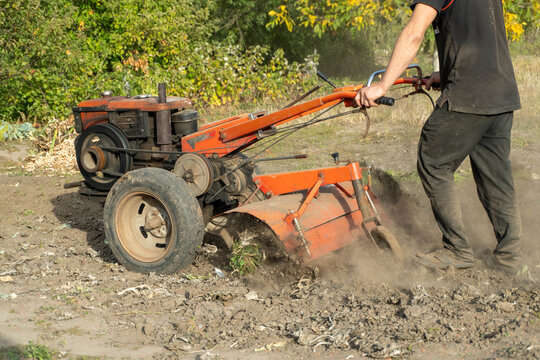 Man operates rotary tiller across rough garden soil, loosening earth beneath its metal hood. Focused farmer guides orange cultivator handles while dirt scatters around working machine.