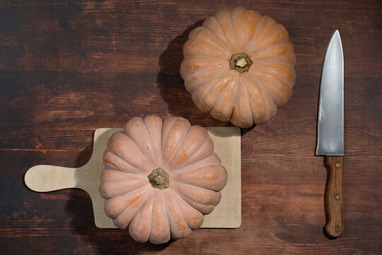 A close-up of two nutmeg squash lying on a wooden table. The squash are photographed from above. A knife lies next to them.