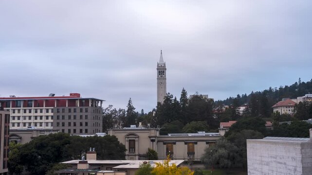 An elevated daytime view of the UC Berkeley campus, featuring Sather Tower (The Campanile) rising above surrounding buildings and trees under a cloudy sky.
