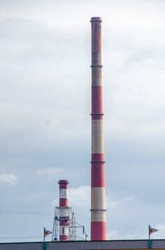 A tall industrial chimney marked with red and white rings rises into an overcast sky. A second smaller stack, power lines and a flag stand below in the background. The view shows heavy industry