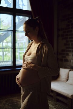 In a cozy indoor setting, a pregnant woman gently cradles her baby bump, lost in thought. Soft natural light streams in through a window, creating a serene atmosphere that enhances the moment