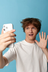 A young man in a white t-shirt takes a selfie, waves at the camera against a solid blue backdrop, conveying casual online greetings and connection. © SHOTPRIME STUDIO