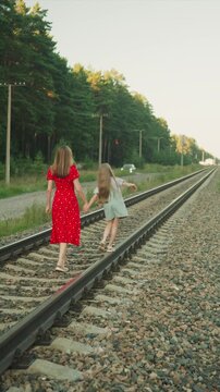 Two young women hold hands as they stroll down parallel railroad tracks. One wears red dress while other is in casual attire with green trees and distant cars visible