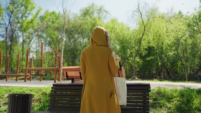 Woman in yellow hooded cardigan approaches and sits on bench carrying art supply bag with wooden handles. scene is set outdoors in park like environment with green trees and other benches