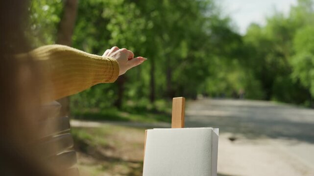 Woman arm extends with yellow sweater preparing to draw on empty white canvas set up outdoors. Bright green trees and pathway visible as background. Sunlight creates soft lens flare