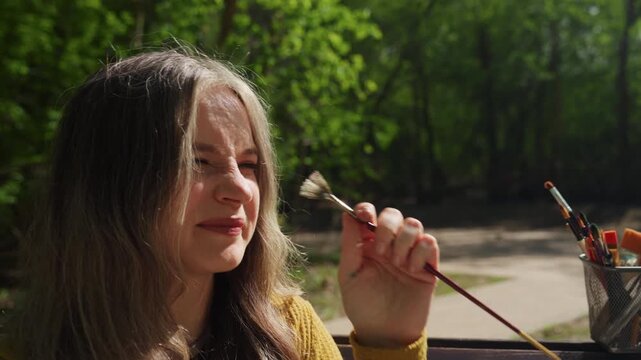 Woman reaches for paintbrush near watercolor set and brush holder in outdoor park setting. Woman holds tool while surrounded by artistic materials with green foliage visible behind her