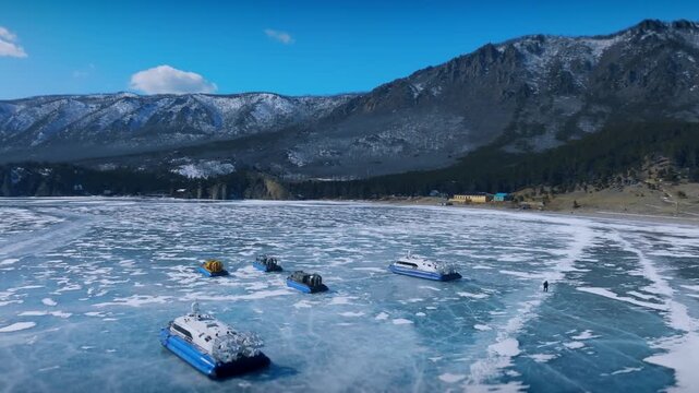Aerial footage shows amphibious hovercraft vehicles resting on the blue ice surface of Lake Baikal in Irkutsk Oblast, Russia under a sunny sky with distant mountain ranges