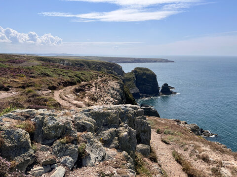 Anglesey, Wales: A rocky coastal cliff path runs through heather and gorse beside steep cliffs, with a sea stack and rock outcrops rising from calm blue water under a partly cloudy sky.