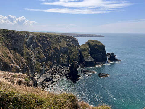 Anglesey, Wales: Steep rocky sea cliffs covered in grass and moss drop to a blue sea, with sea stacks and rocks at the base, a distant headland on the horizon and wispy clouds in a blue sky.