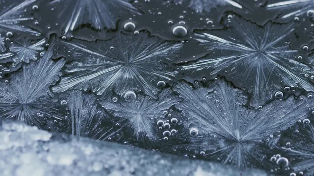 Ice crystals form delicate star shapes on a cold surface. Bubbles float nearby, adding texture to the frozen scene. Snowy patterns shimmer under dim, cool light