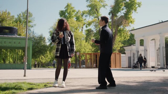 Young woman holds her phone in outdoor park setting as another woman walks towards her. scene includes greenery paved path and distant people walking or cycling under bright daylight