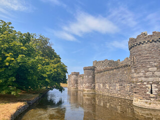 Beaumaris Castle, Isle of Anglesey, - july 17, 2026: Stone curtain walls and round towers of a medieval castle rise beside a water-filled moat, with a large leafy tree on the bank under a blue sky. © AnyaWhy