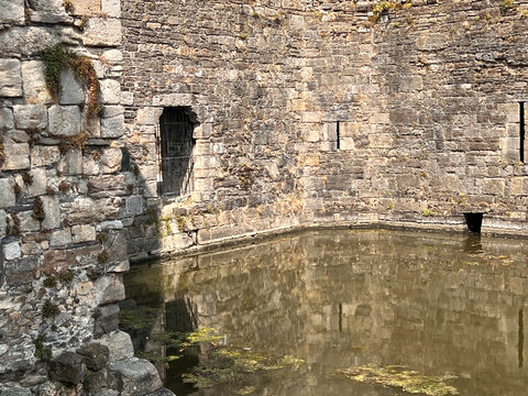 Beaumaris Castle, Isle of Anglesey, - july 17, 2026: Weathered stone castle wall with a barred window and narrow arrow slits rises from a still, algae-dotted moat reflecting the masonry.