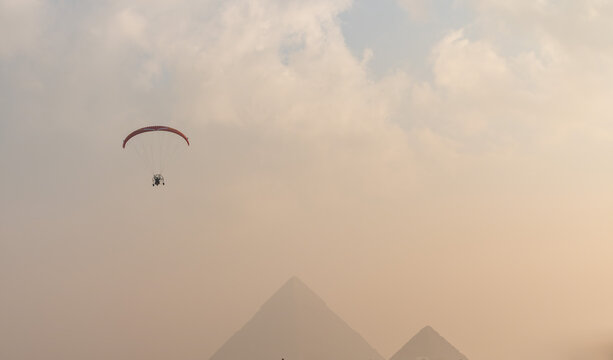 View of a paramotor flying over the Giza Necropolis with the ancient pyramids visible through the hazy sky in Al Haram, Giza Governorate, Egypt.