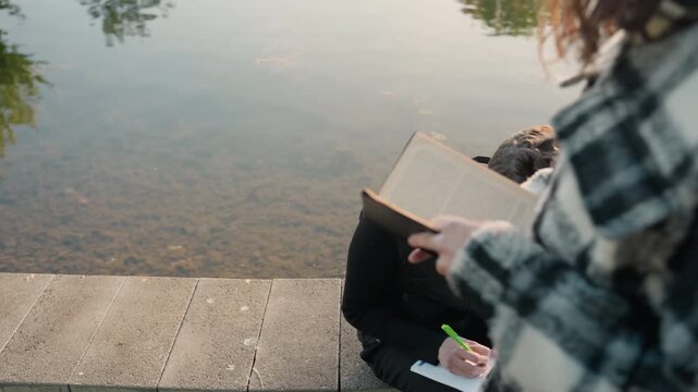 Person writes in notebook by hand while another holds open book and smartphone near with calm water reflections writing pen paper text study education