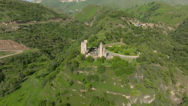 Scenic aerial footage captures the historic stone towers of Vatan memorial complex surrounded by lush green hills, steep ridges, and valleys in Dagestan, Russia on a sunny day