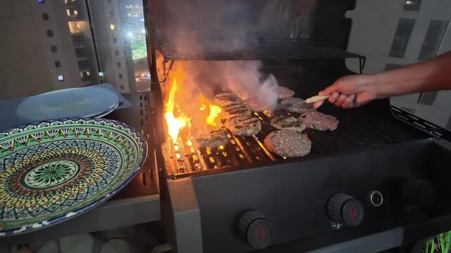 Burgers grilling on an outdoor gas barbecue with open flames and rising smoke, as a hand turns the patties with a spatula beside a patterned serving plate