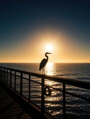 Fototapeta premium Silhouette of great blue heron standing on wooden pier at sunset. Large wild bird watches ocean waves under bright golden sun. Peaceful coastal nature scene with calm sea, backlight sun rays during