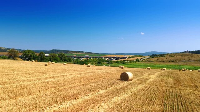 Round straw bales across a field in Slovakia. A cylindrical bale stands in the foreground of cut farmland, with a distant bridge and wooded hills.