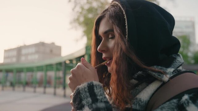 Woman with long hair wears black hooded jacket and carries brown strap bag. Woman holds object near her mouth in soft natural light with green structures and distant buildings visible