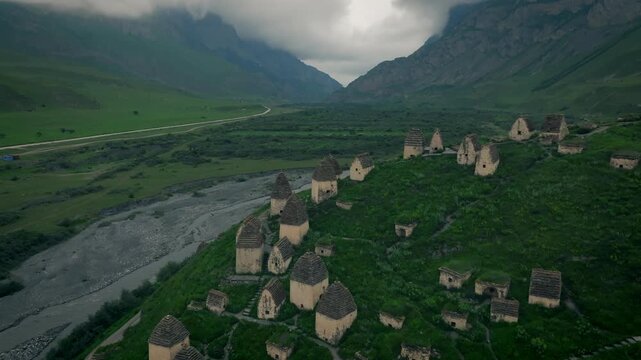 High angle aerial view of historic medieval stone crypts at Dargavs village necropolis surrounded by green mountain valleys and riverbed in North Ossetia-Alania, Russia