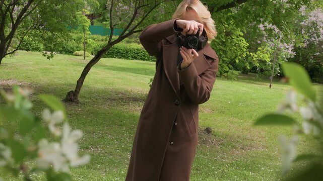 Woman holding black camera taking pictures outdoors. Brown coat visible. Green trees and grass background lady photographer outdoor nature greenery park