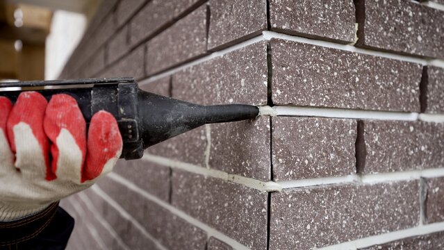 Red work glove holding caulking gun applying white sealant to brown brick wall corner, exterior facade finishing