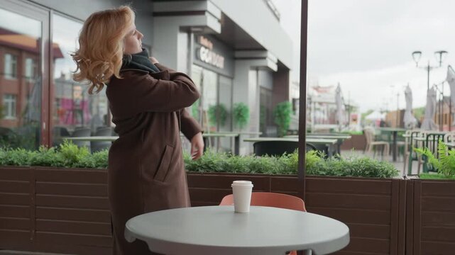 Woman sits at outdoor cafe table with coffee cup. Woman stands up picks up cup from round gray table on orange chair and leaves coat scarf urban street