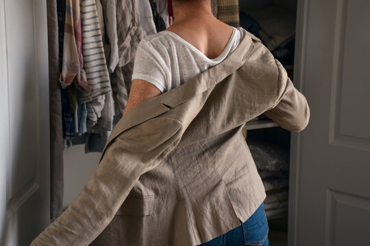 Back view of a woman putting on or taking off a beige linen blazer over a white t-shirt in front of an open closet