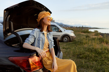 Naklejka premium A relaxed woman in a denim jacket and dress sits on a car trunk by a coastal meadow, enjoying warm sunlight. A lifestyle outdoor fashion casual portrait concept, serene and cheerful.